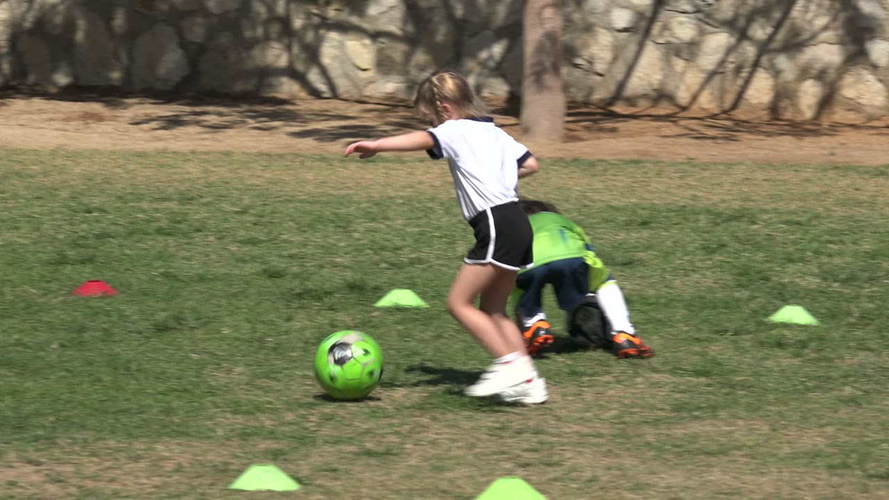 Girl runs beside a green soccer ball as another child falls on the grass.