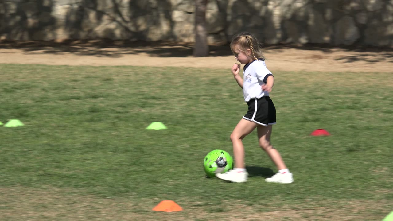 In Los Cabos, Mexico, the girl runs in profile near a bright green soccer ball, with green and orange cones spread across the sunlit grass and a bench in the background.
