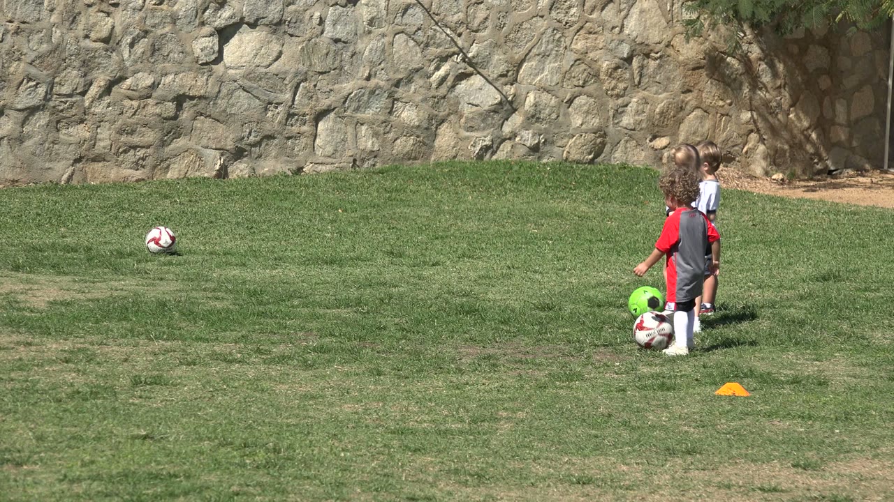 Two children in soccer clothing move near a white ball and a green ball on the Los Cabos grass field, with an orange cone marking the drill area.