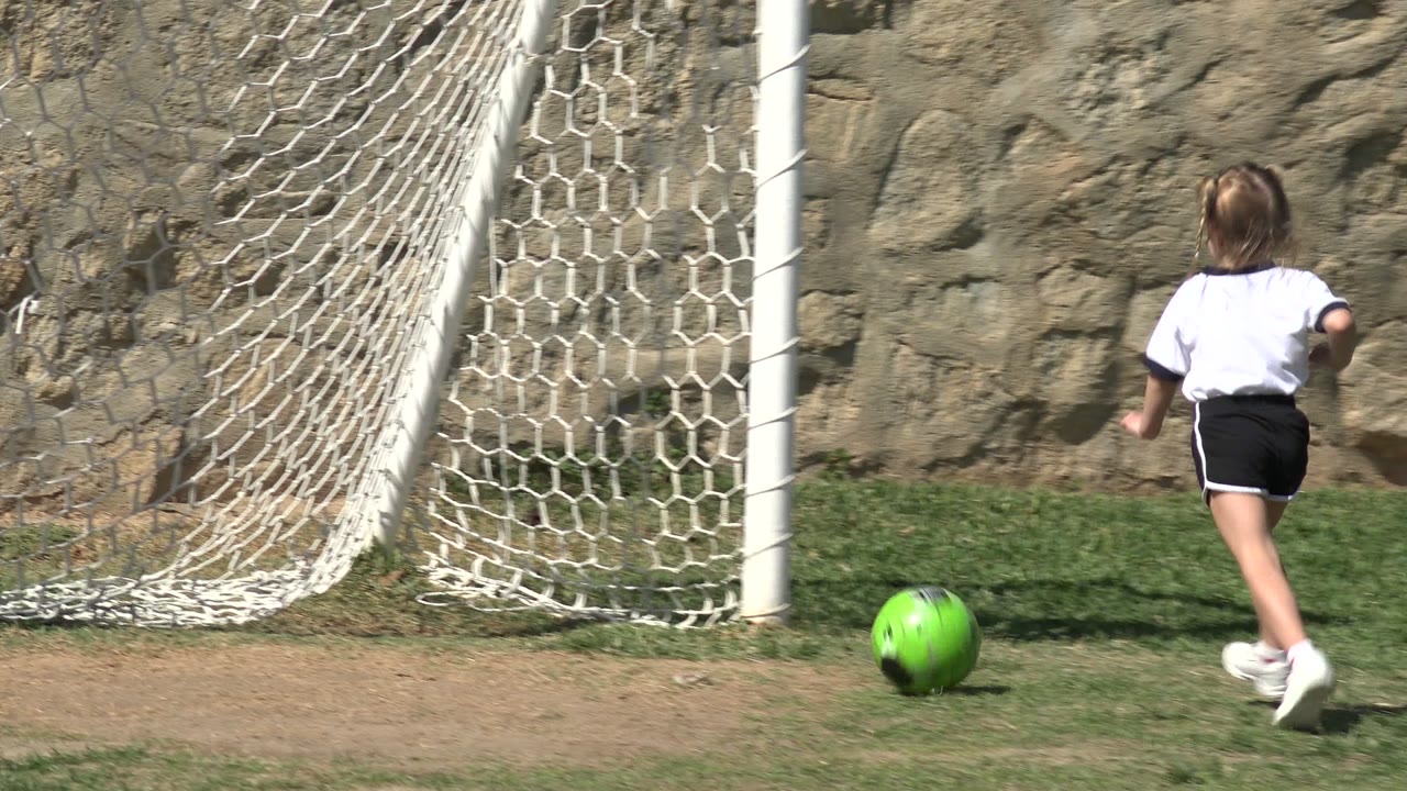 Young girl runs toward a green soccer ball beside a small goal.
