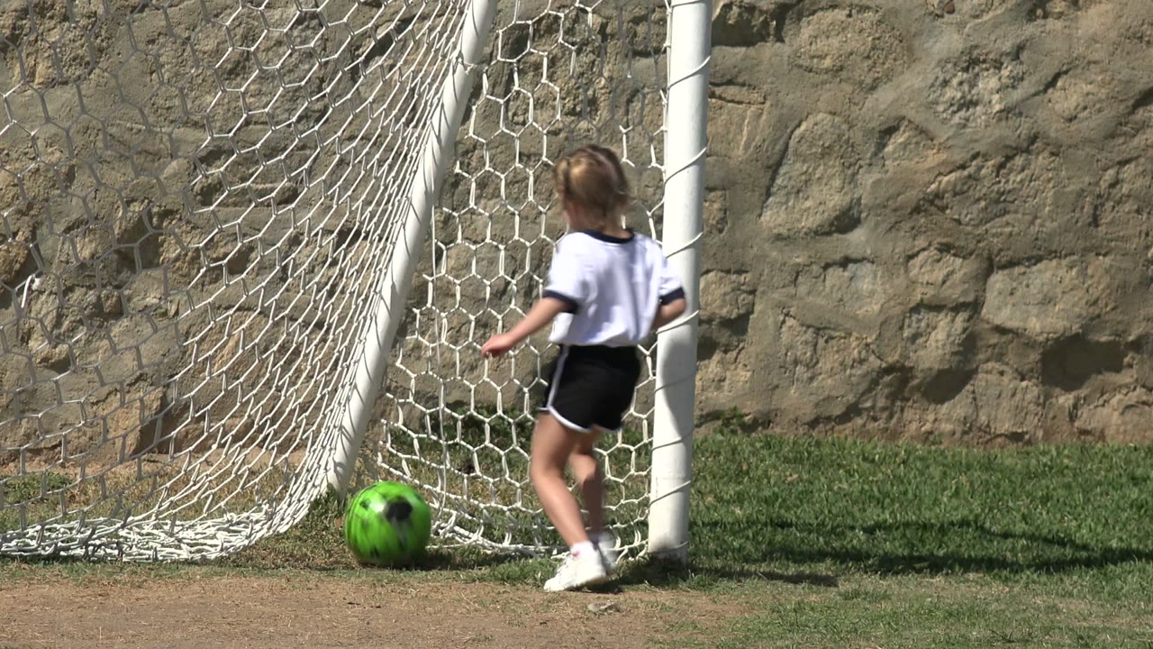 The girl follows the bright green soccer ball as it reaches the white net, with the stone wall and grass field behind her in Los Cabos, Mexico.