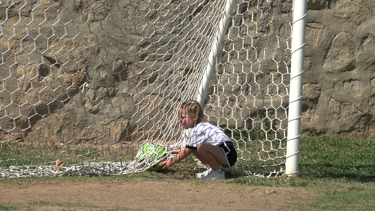 Girl crouching inside a soccer goal with a green ball.