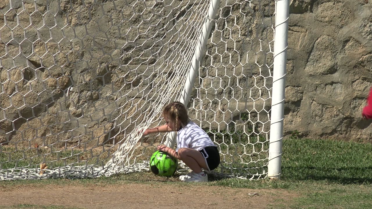 The girl crouches near the white goalpost in Los Cabos, Mexico, with one hand near the green ball and the net hanging against the stone wall.