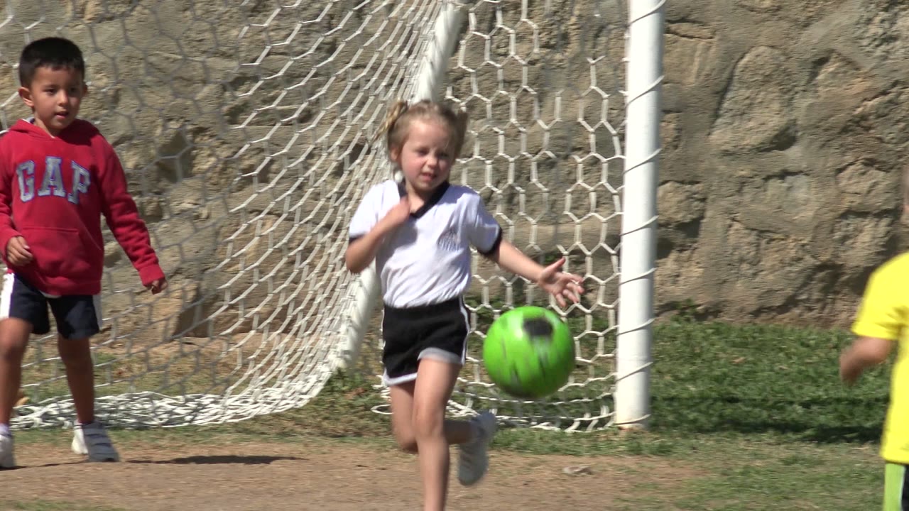 A young girl in a white soccer shirt runs toward a bright green soccer ball near a white goal net in Los Cabos, Mexico, with another child in a red sweatshirt behind her.