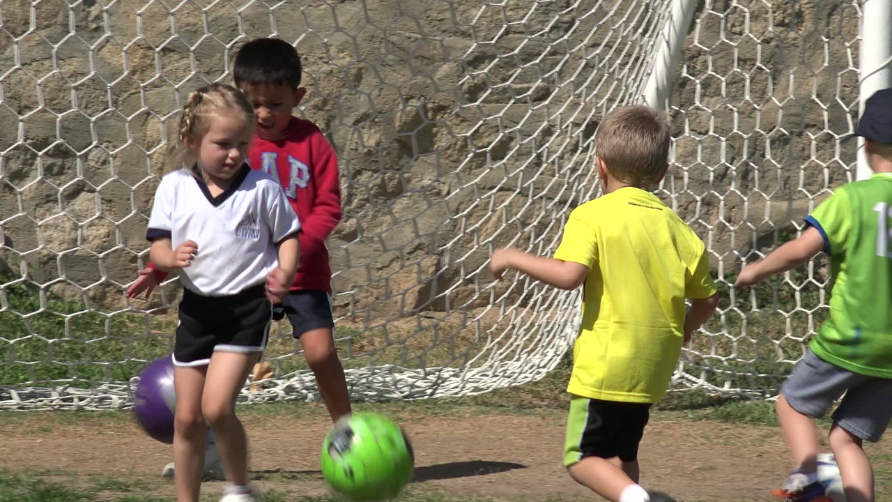 A girl in a white jersey and a boy in a red sweatshirt stand near green and purple soccer balls beside the goal net in Los Cabos, Mexico.