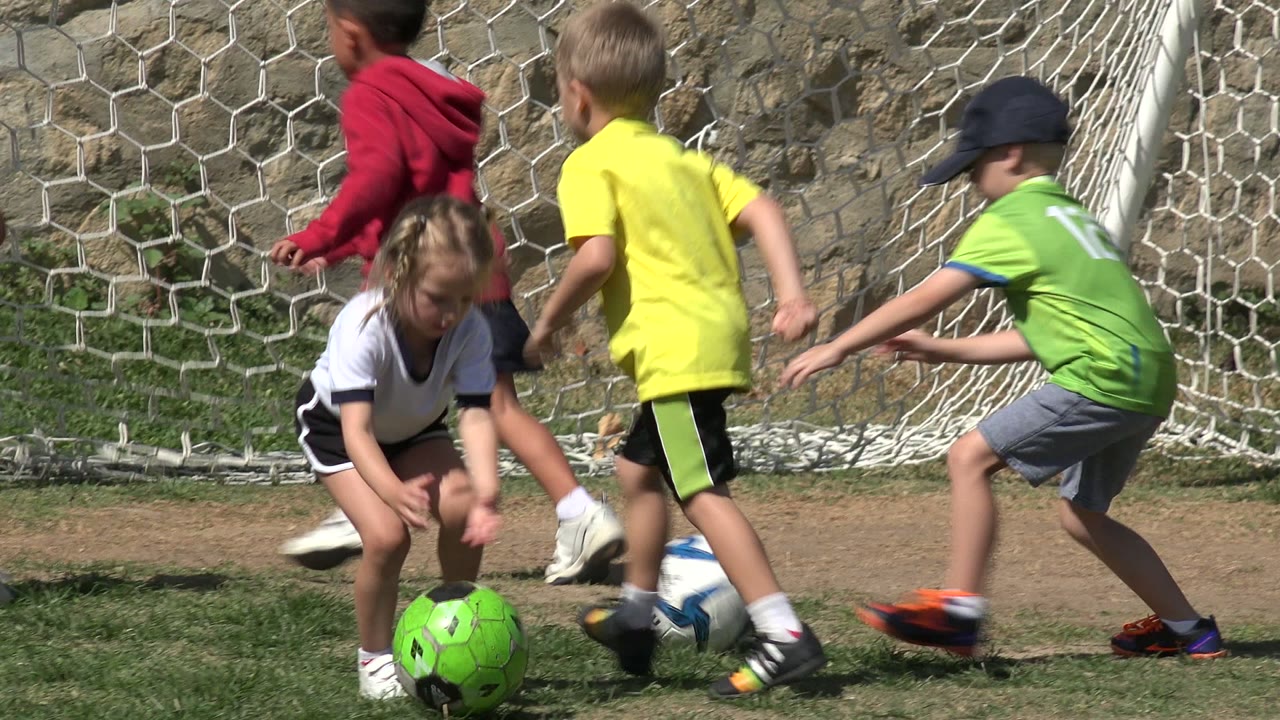 Children in yellow and green jerseys chase a soccer ball near the goal.