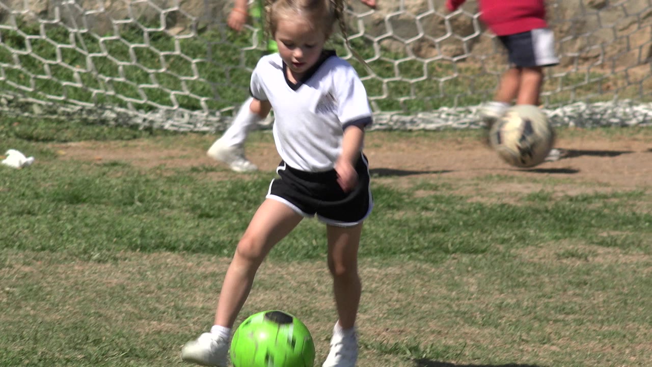 The girl moves her foot beside the bright green soccer ball on the Los Cabos grass field while other children are partly visible near the goal net.