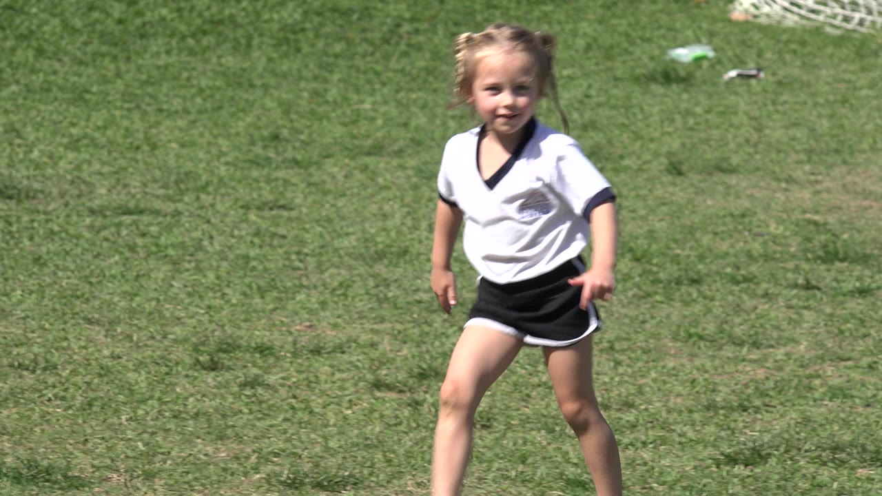 Young blonde girl in a white soccer jersey and black shorts faces the camera on a sunny grass field in Los Cabos, Mexico.