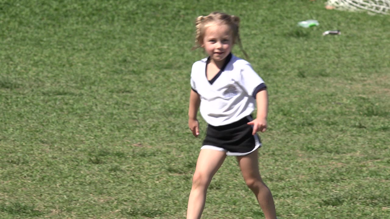 The girl remains near center frame in her white and black soccer uniform, standing on the grass field in Los Cabos, Mexico.