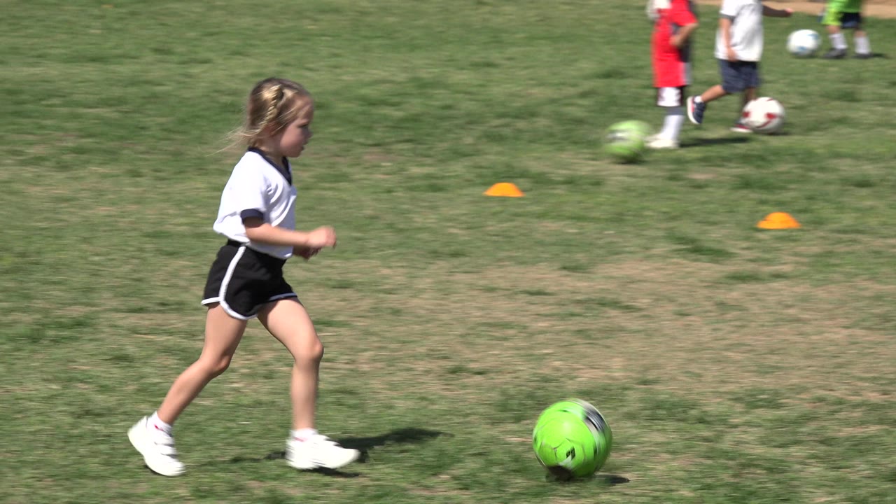 The child jogs across patchy grass near the green soccer ball, with orange cones and several young soccer players blurred in the background in Los Cabos, Mexico.