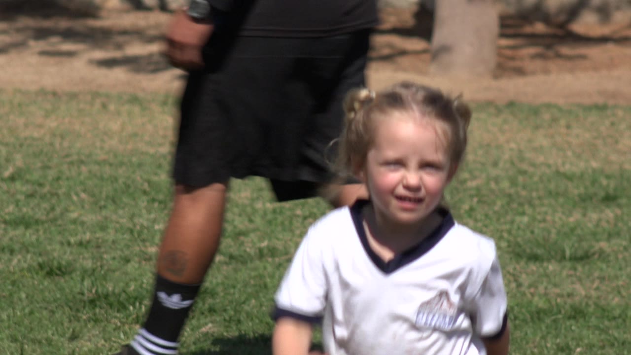 The young girl remains centered in a white shirt on the Los Cabos soccer field, with patchy grass and an adult passing in the background.