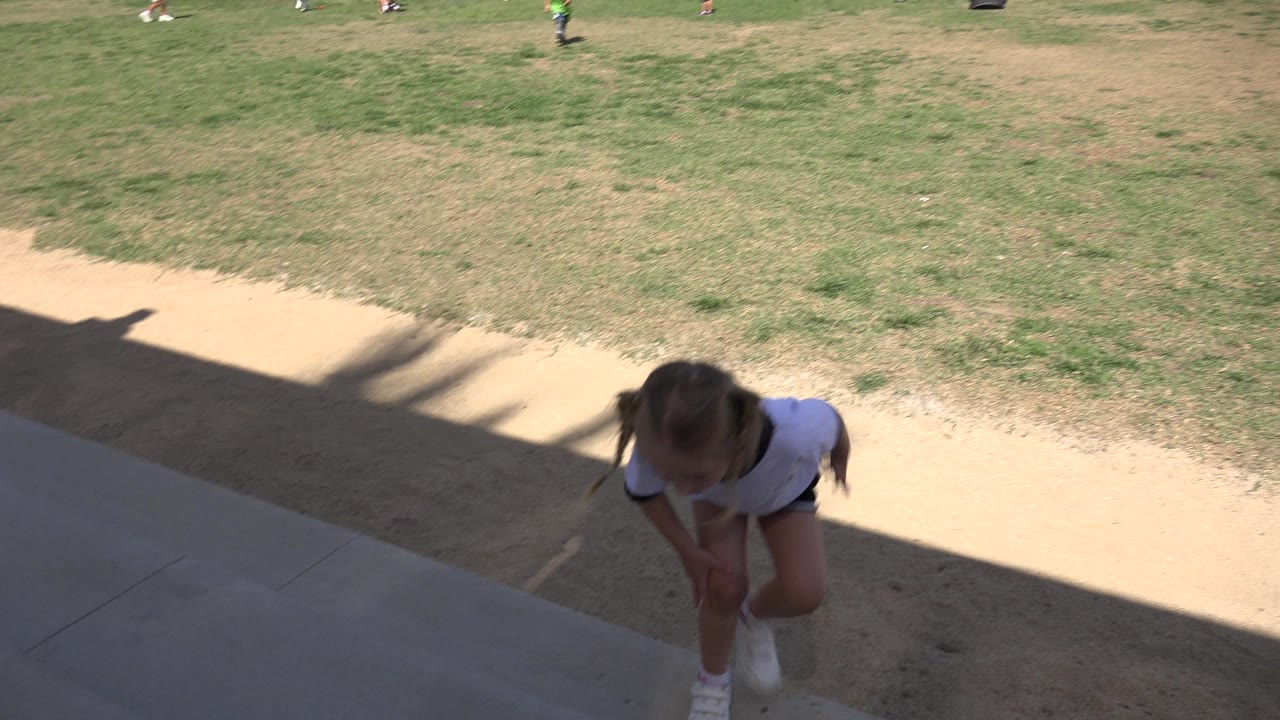 Young girl in soccer clothes steps up from a shaded sideline near a grassy field.
