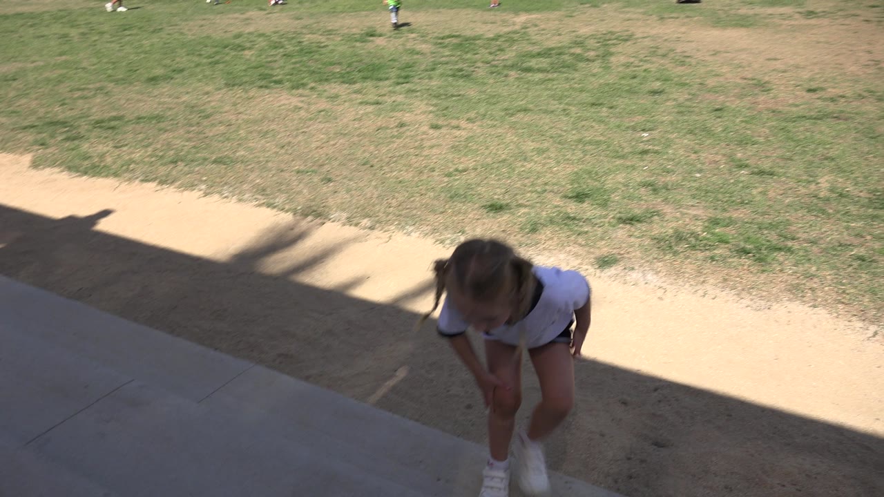 The girl bends forward near the sideline shade in Los Cabos, Mexico, while a dry grassy soccer field and small background players fill the upper frame.