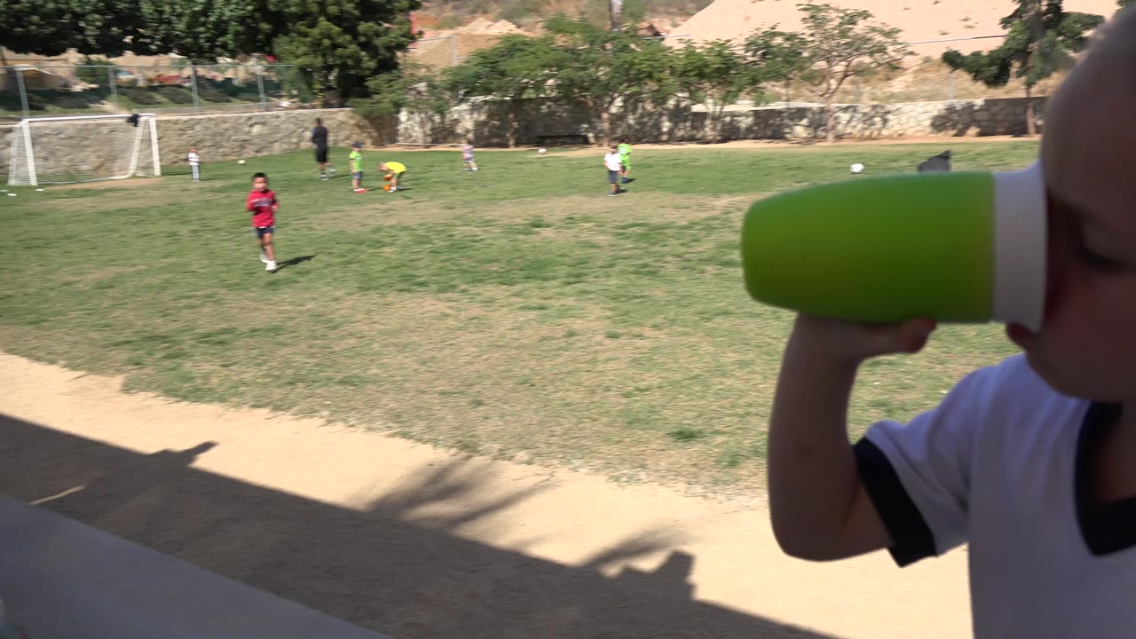The girl keeps the green cup to her mouth near the right edge of the frame as small soccer players move across the Los Cabos park field behind her.