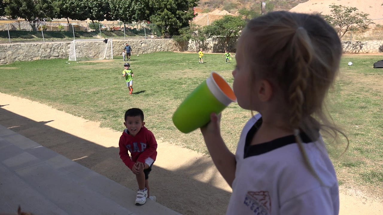 A child drinks from a green cup close to the camera as a boy in a red GAP hoodie approaches from the sunlit grass field in Los Cabos, Mexico.