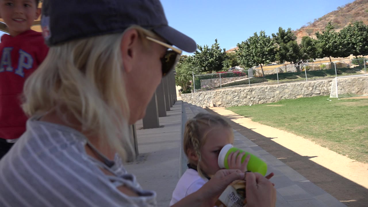 In Los Cabos, Mexico, the woman remains in close foreground profile while the girl raises a green cup near her face beside the shaded paving and bright grass.