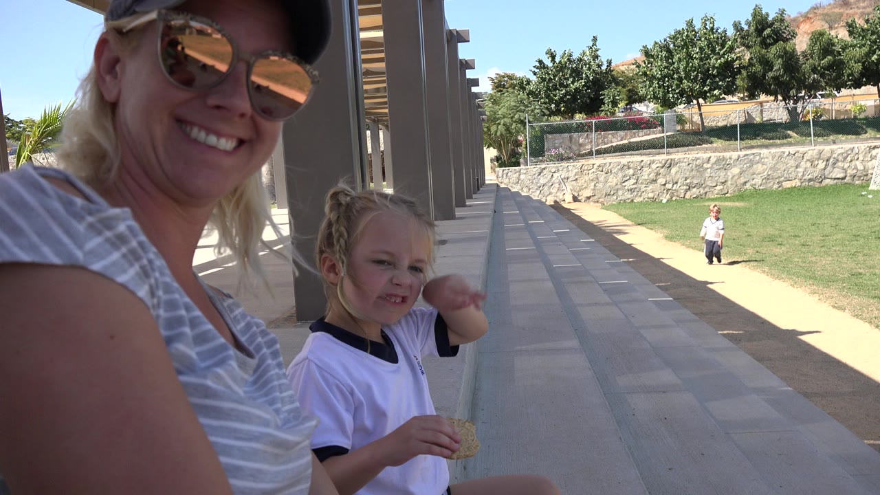 Woman in cap and sunglasses beside a young girl under a shaded outdoor structure.