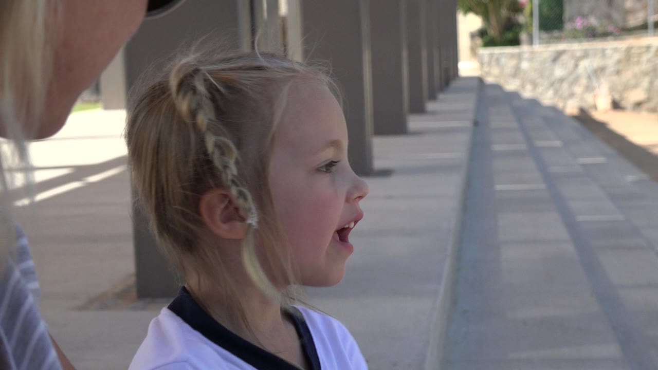 A young blonde girl with braided hair sits beside a partially visible adult under a shaded walkway in Los Cabos, Mexico, with columns and a bright path behind her.