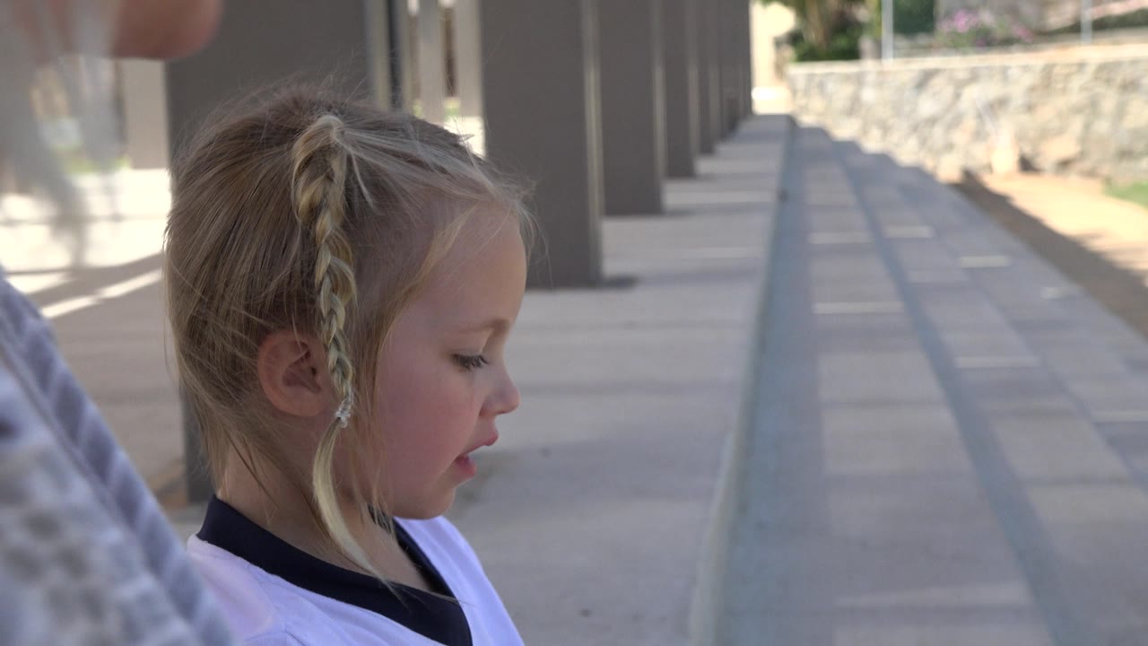 Girl looking downward beside an adult on a shaded walkway.