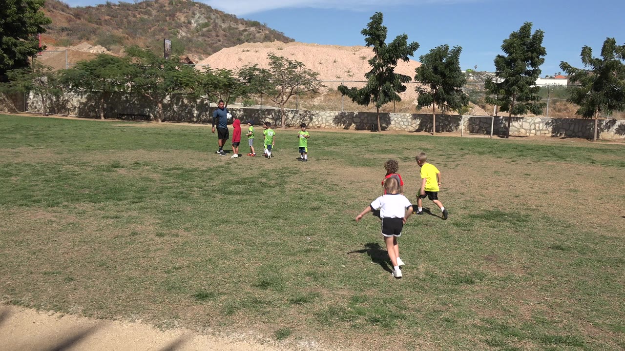 Wide view in Los Cabos, Mexico of children running across a patchy grass field toward an adult coach and a small group of kids near trees and dry hills.