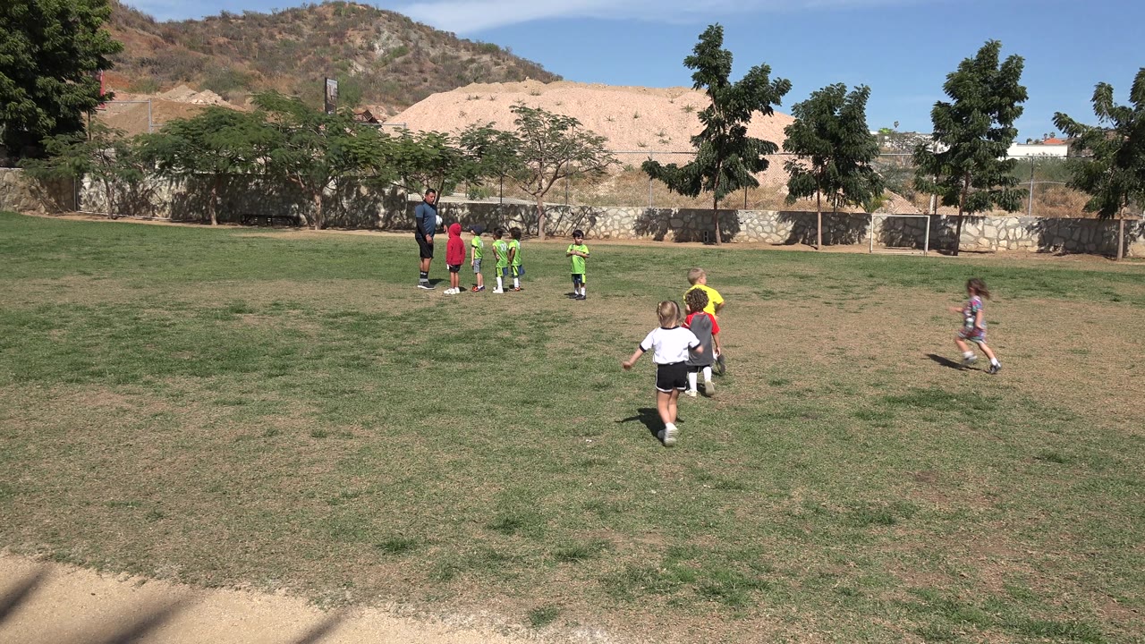 A wider Los Cabos, Mexico field view shows several children moving toward a cluster of kids and an adult coach, with sparse trees and dry hillside terrain behind the fence.
