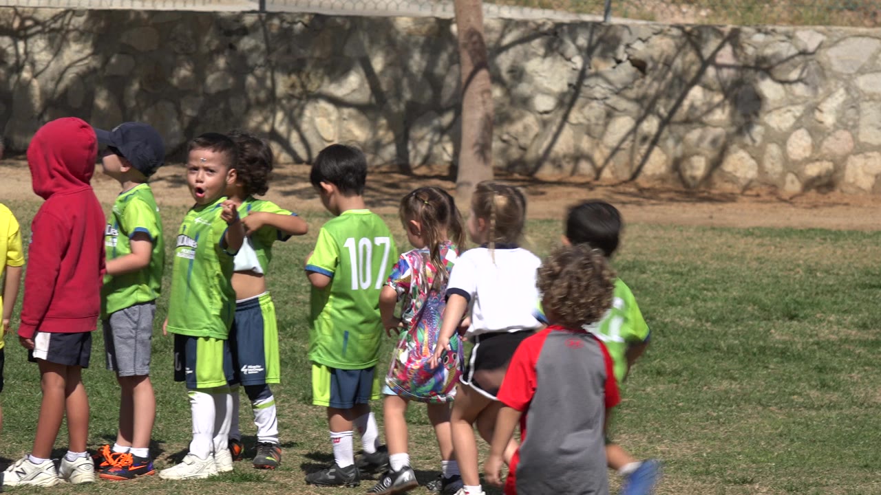The group remains gathered on the grass in Los Cabos, Mexico, with jersey numbers visible on green uniforms and several children facing different directions.