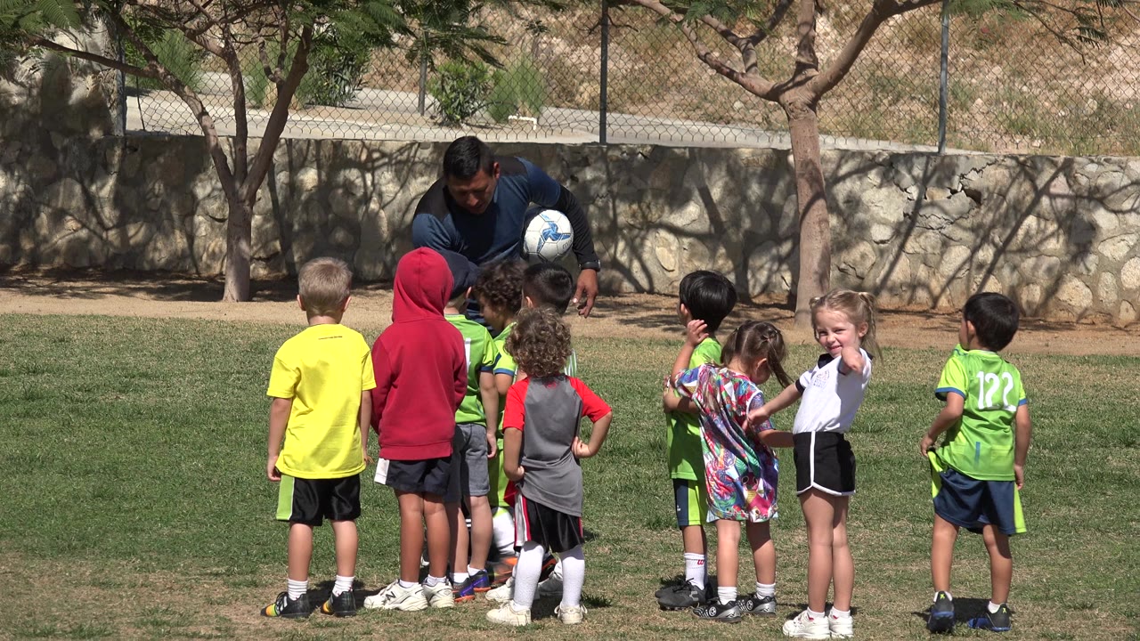 At 0:00 in Los Cabos, Mexico, a coach holding a soccer ball bends toward a group of small children in bright jerseys on a grassy field.