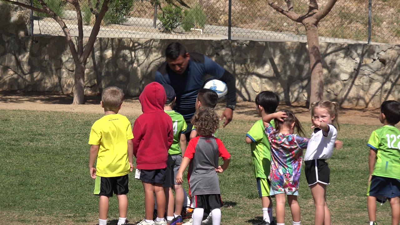 Coach with soccer ball speaks to children on grass.