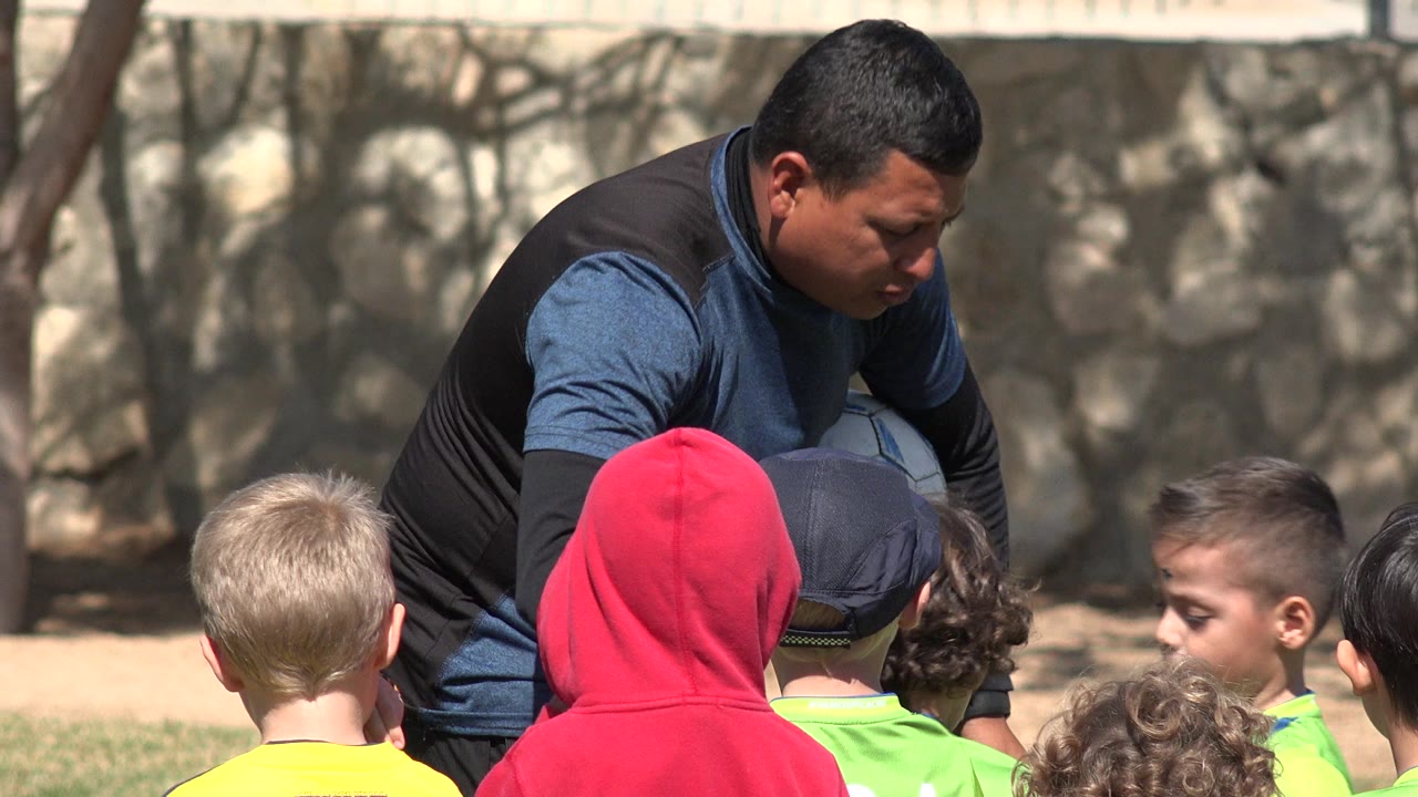 In Los Cabos, Mexico, a coach crouches with a soccer ball partly visible while children in bright shirts listen from the foreground near a stone wall and fence.