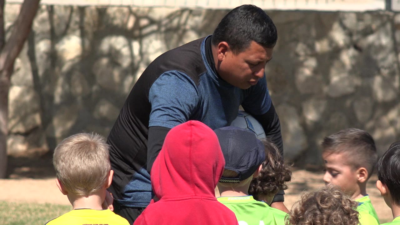 The coach remains crouched in Los Cabos, Mexico, with children facing him from the foreground and a soccer ball tucked against his body in bright outdoor light.