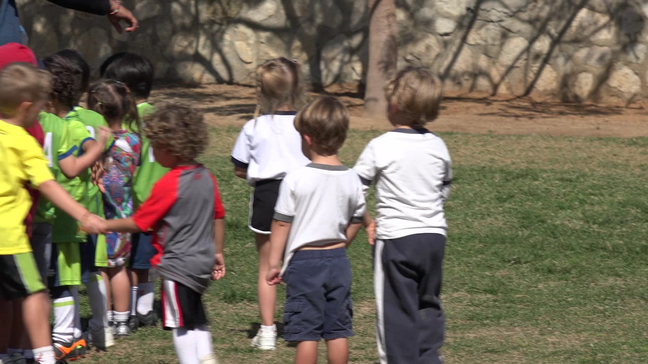 Children in sportswear stand on a grass field near a coach's cropped arm.