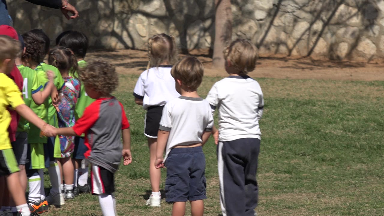 Children in green uniforms and white shirts cluster on a sunlit grass field in Los Cabos, Mexico, with a cropped adult figure standing nearby.
