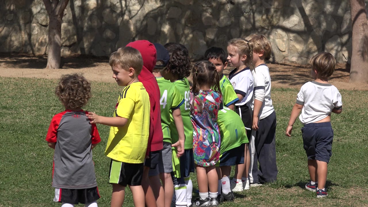 The children remain grouped on the grass in Los Cabos, Mexico, with green jerseys, a yellow shirt, and white tops visible against the shaded tree and wall background.