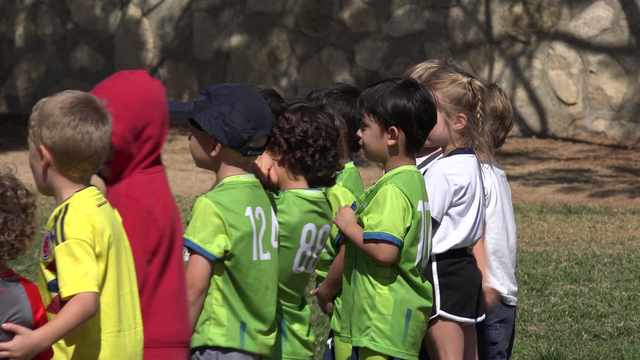 The group of children remains packed together on the grass in Los Cabos, Mexico, with green jerseys most visible and a blonde girl in a white shirt near the back.