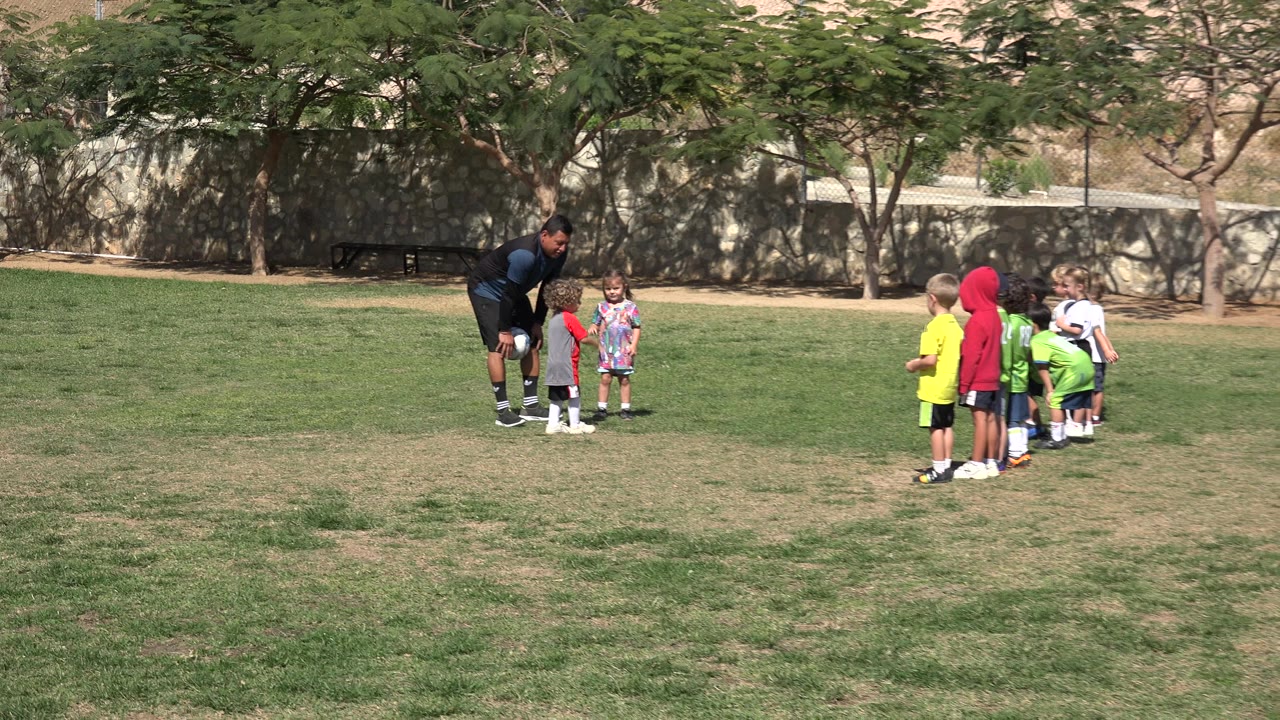 In Los Cabos, Mexico, the adult remains bent near a child with a ball visible by his side as other children face the interaction from the right side of the field.