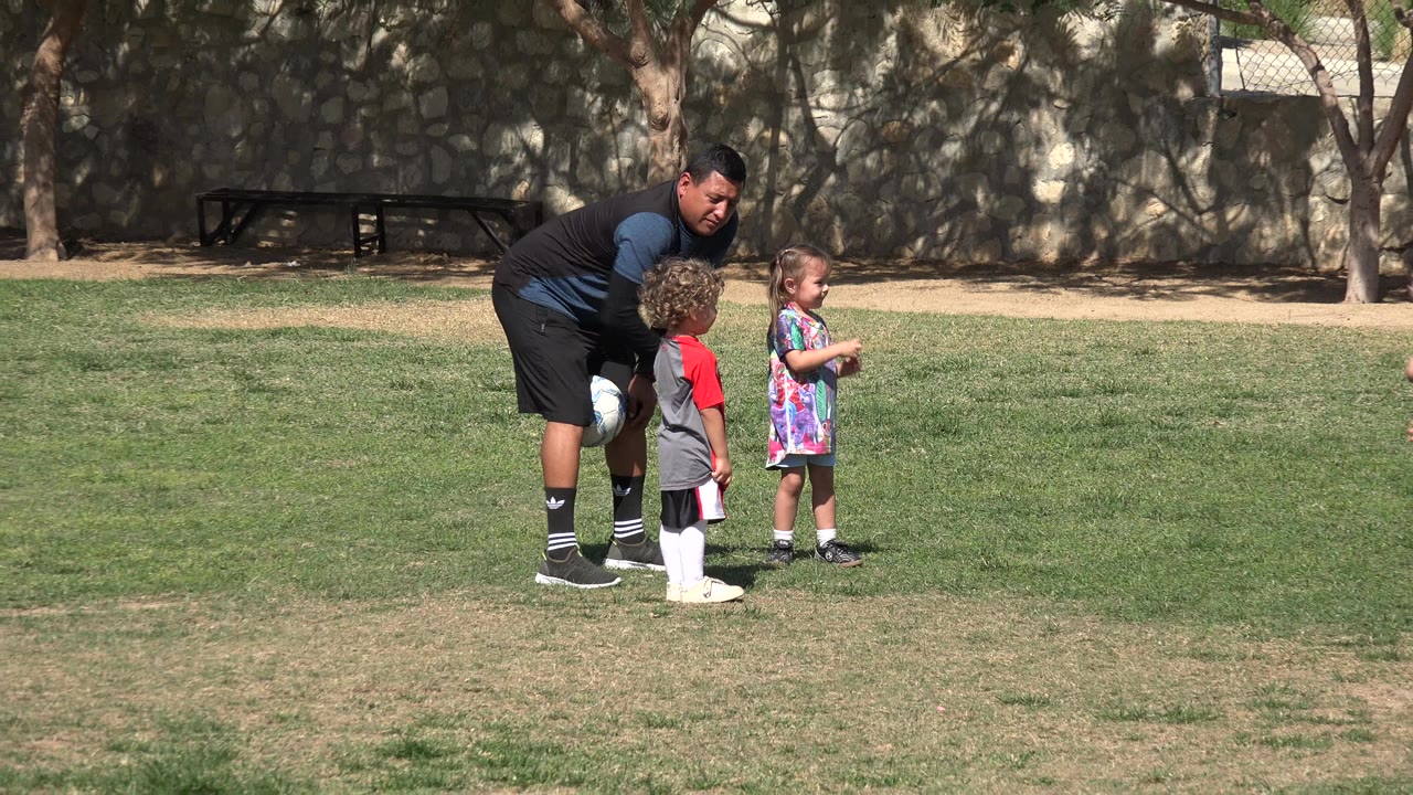 The group remains on the grass in Los Cabos, Mexico, with the adult leaning toward a child while another child stands nearby in bright clothing.