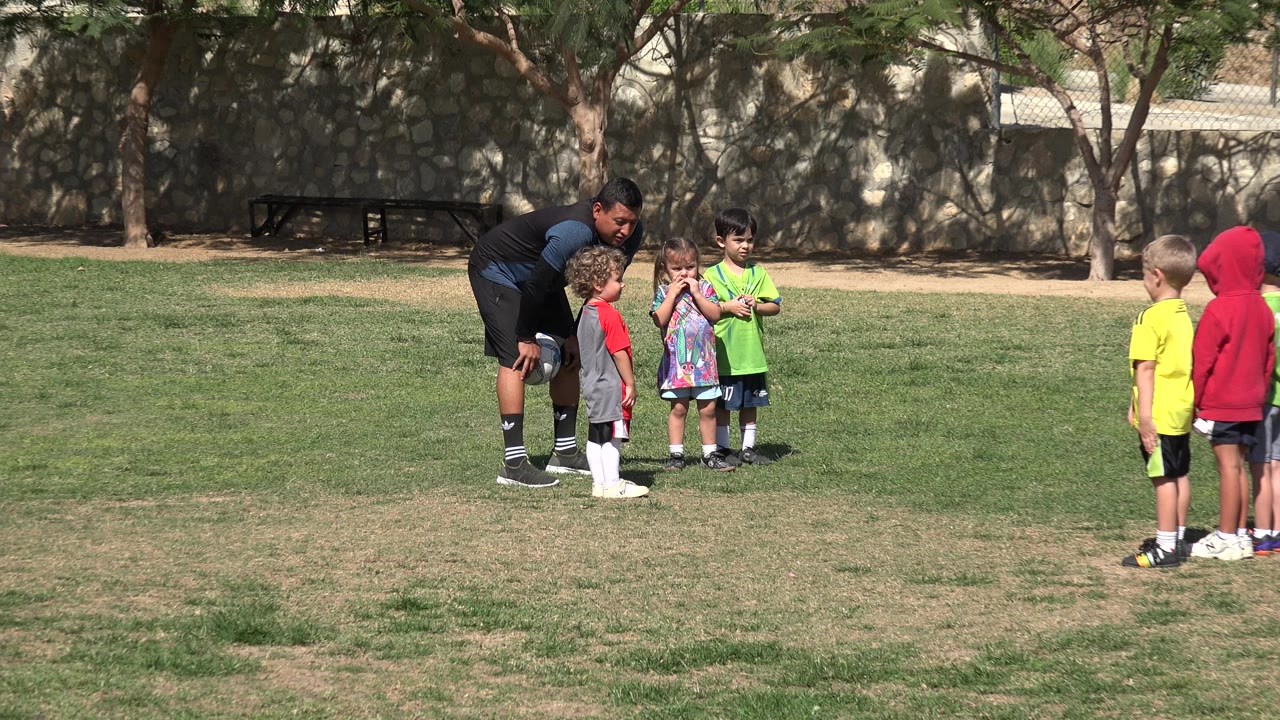 The coach remains bent toward several children in Los Cabos, Mexico, as two young players in yellow and red stand apart on the grass.