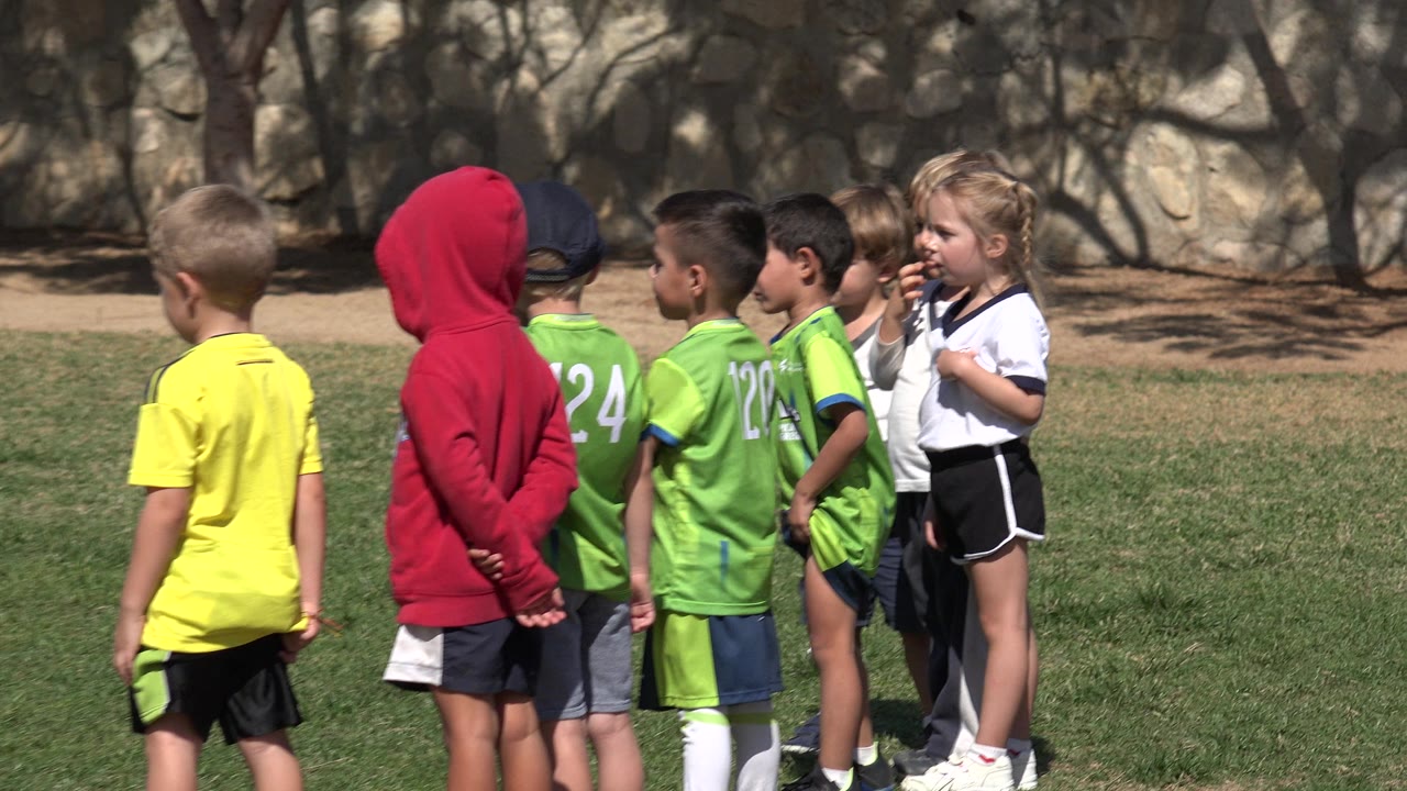 A youth soccer group stands in bright sun on a field in Los Cabos, Mexico, with several green jerseys facing a child in a white-and-black uniform.