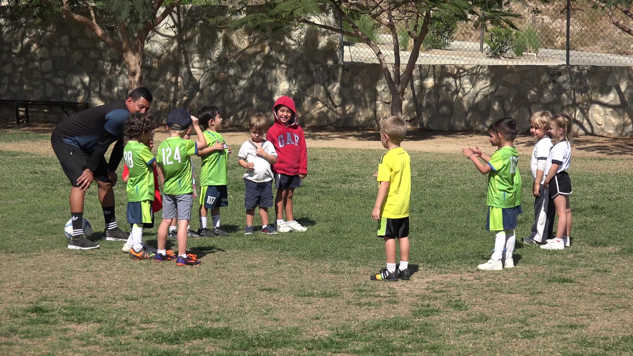 Children in green and white football uniforms stand near an adult coach on a grass field in Los Cabos, Mexico, with trees and a stone wall behind them.
