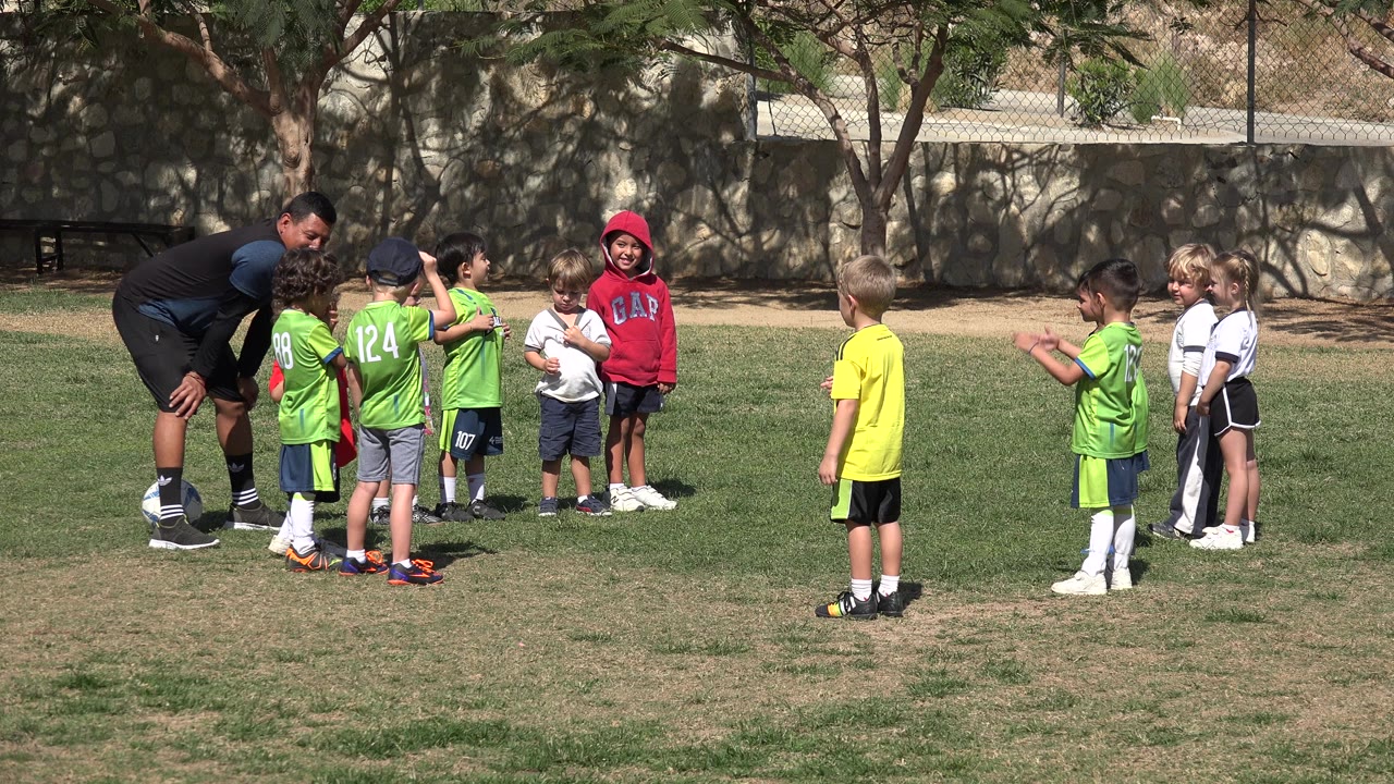 A coach bends toward children in green jerseys while other children in white kits wait to the right on the Los Cabos grass field.