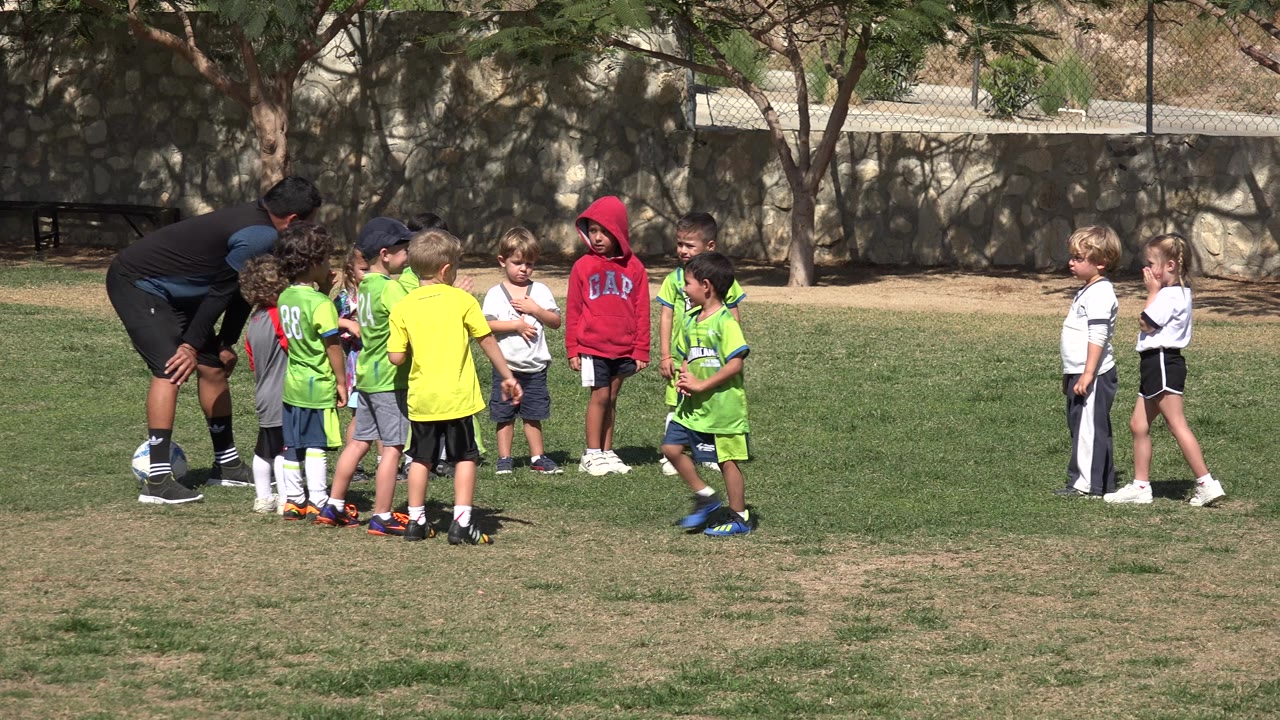 The youth soccer group remains clustered around the coach in Los Cabos, Mexico, with green jerseys facing inward and two children in white waiting on the right side of the frame.