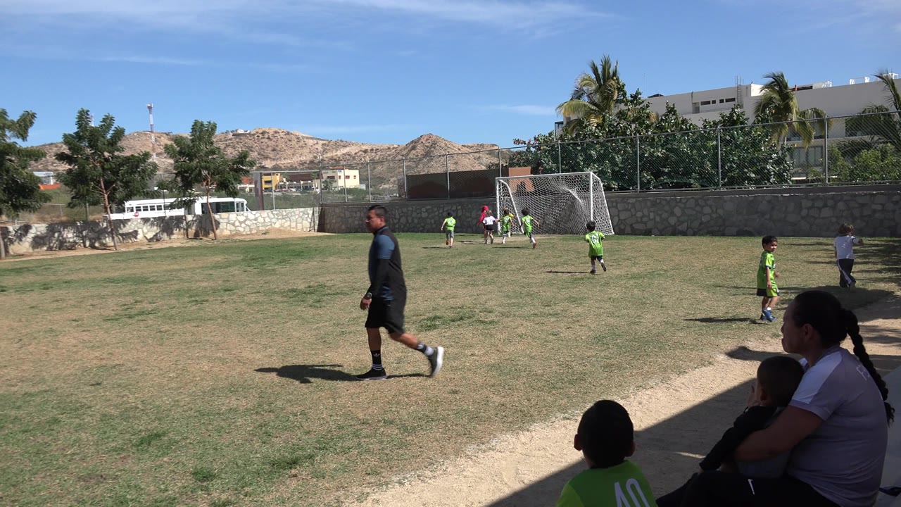 Wide Los Cabos soccer field frame with an adult walking in the foreground, children in green jerseys near the white goal, and spectators sitting at the lower right edge.