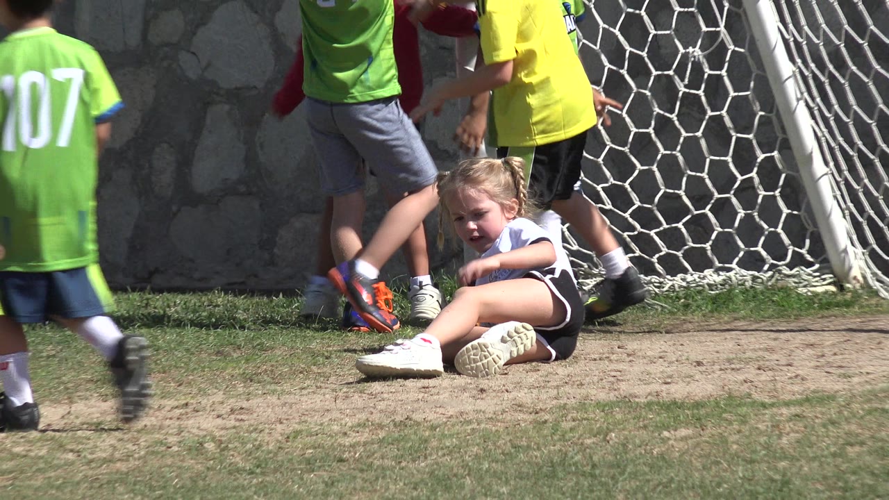Girl sitting on dirt near a soccer goal as children play nearby.