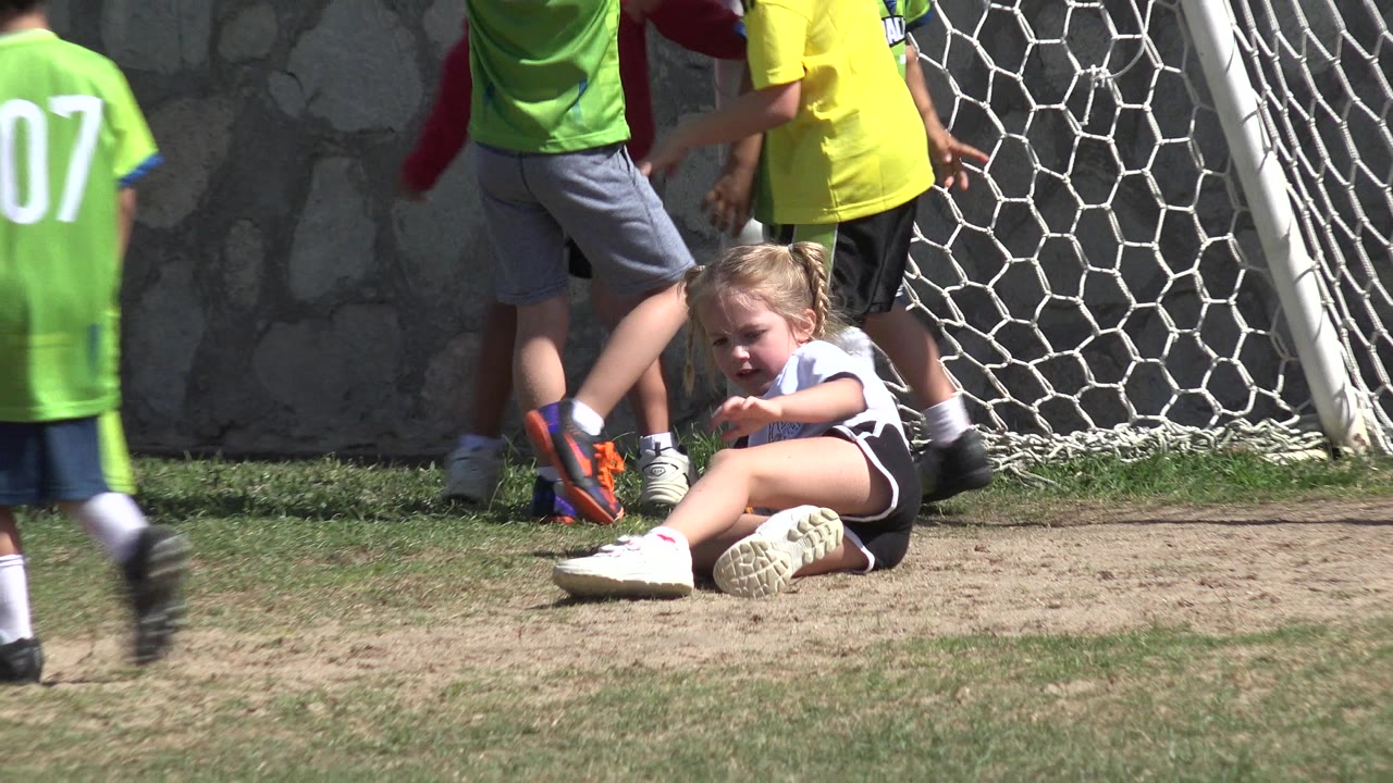 The seated girl remains on the dirt beside the white goal net in Los Cabos, Mexico, with a green-jerseyed child crossing the foreground.