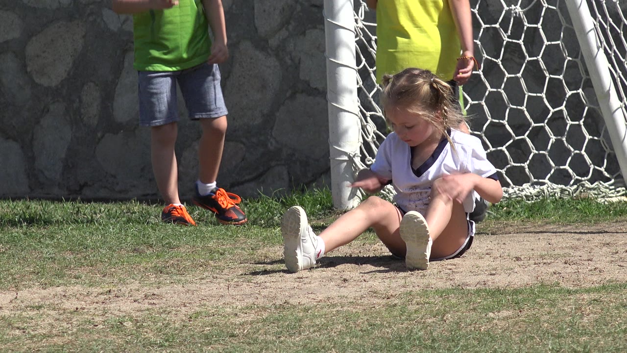 Young girl sitting on grass near a soccer goal with boys standing behind her.