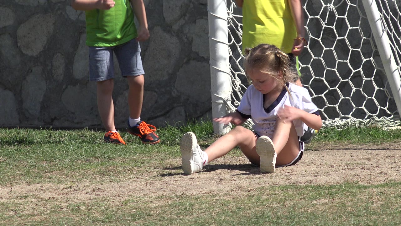 The girl remains seated in front of the Los Cabos goal net, looking downward as boys in green and yellow shirts stand near the white post and stone wall.