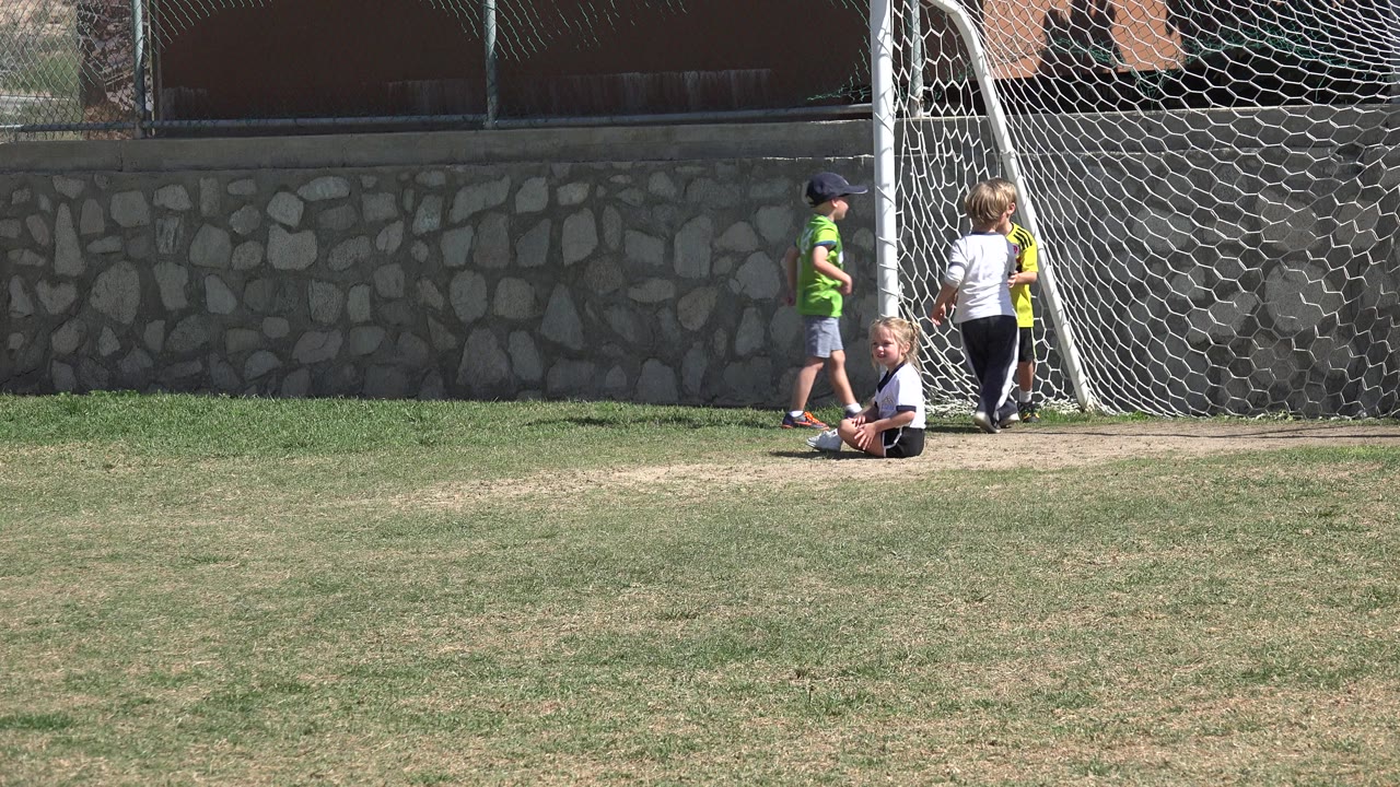 Wide frame in Los Cabos, Mexico showing a girl with pigtails seated near a soccer goal while other children stand by the net against a stone wall.