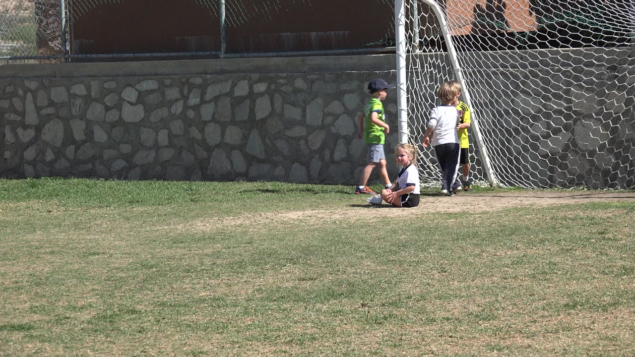 Wide frame in Los Cabos, Mexico with a young girl sitting on the grass in front of the soccer goal as children in green and white shirts stand nearby.