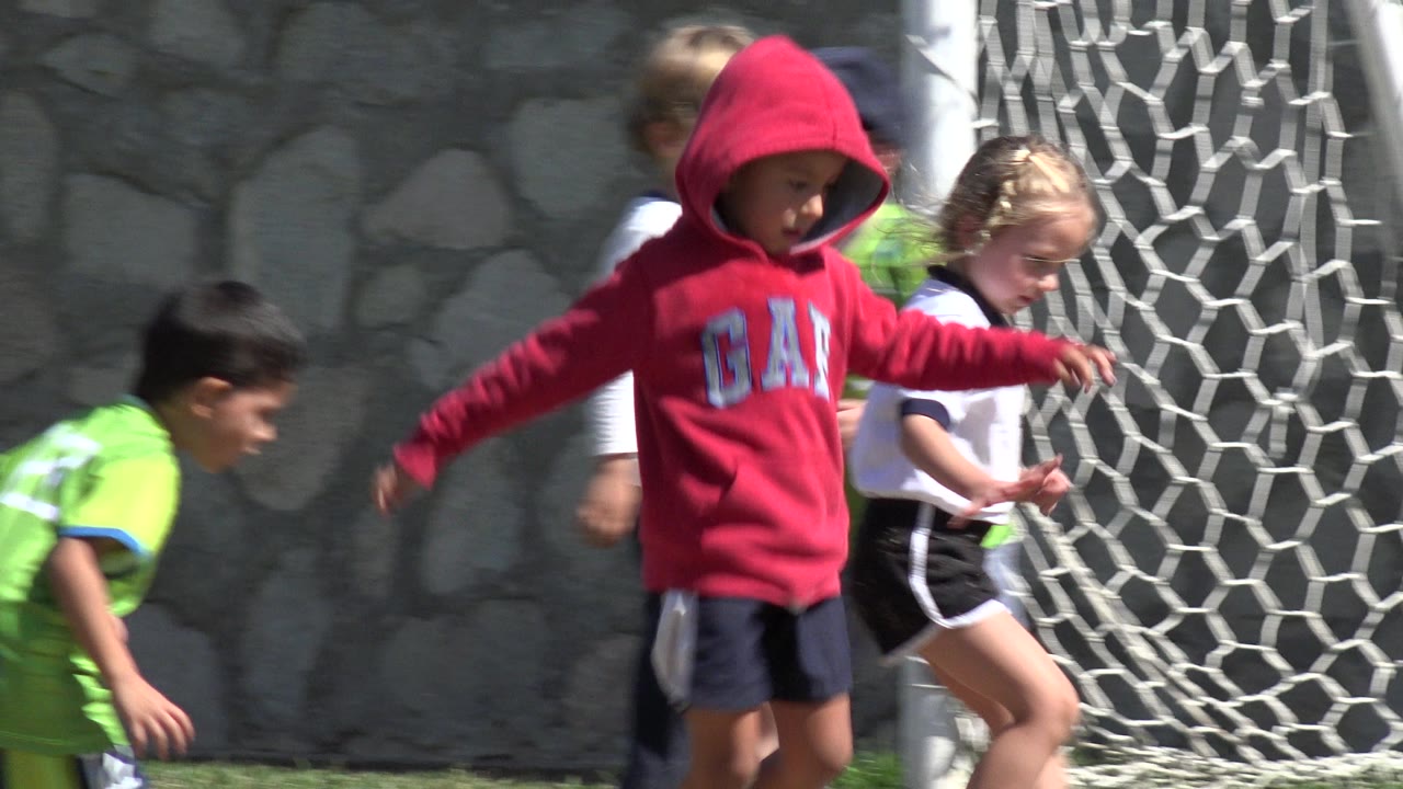 A child in a red hooded sweatshirt and other children move in front of the goal net on a sunlit field in Los Cabos, Mexico.