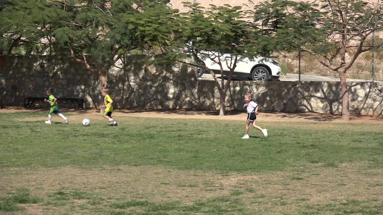 Two children in bright jerseys move near the soccer ball while another child in a white shirt walks across the grass in Los Cabos, Mexico.